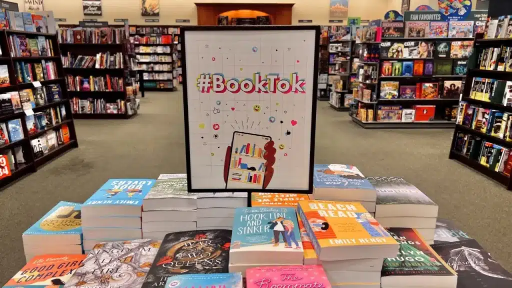 Ein Buchladen mit einem Tisch voller Bücher, auf dem ein Schild mit der Aufschrift "#BookTok" steht. Das Schild zeigt eine Illustration mit einem Smartphone, aus dem kleine Icons und Buchbilder herausspringen. Im Hintergrund sind Regale mit weiteren Büchern und Spielen zu sehen. Die Szene symbolisiert den Einfluss von BookTok auf die Buchbranche.