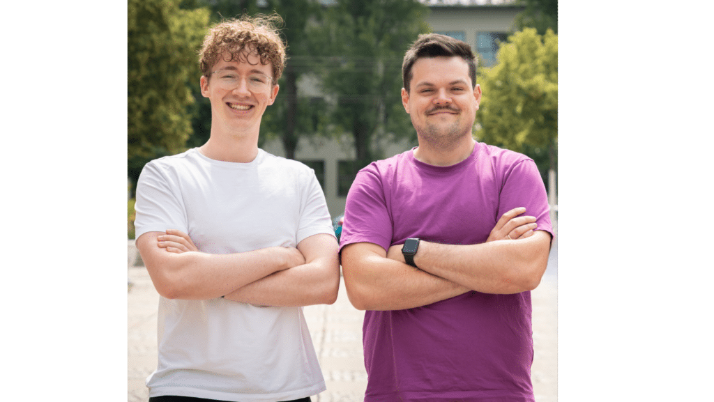 Zwei lächelnde Männer stehen nebeneinander mit verschränkten Armen. Der Mann links trägt ein weißes T-Shirt und eine Brille, während der Mann rechts ein lila T-Shirt und eine Smartwatch trägt. Im Hintergrund sind Bäume und ein Gebäude bei Tageslicht zu sehen. Die Szene wirkt freundlich und professionell.